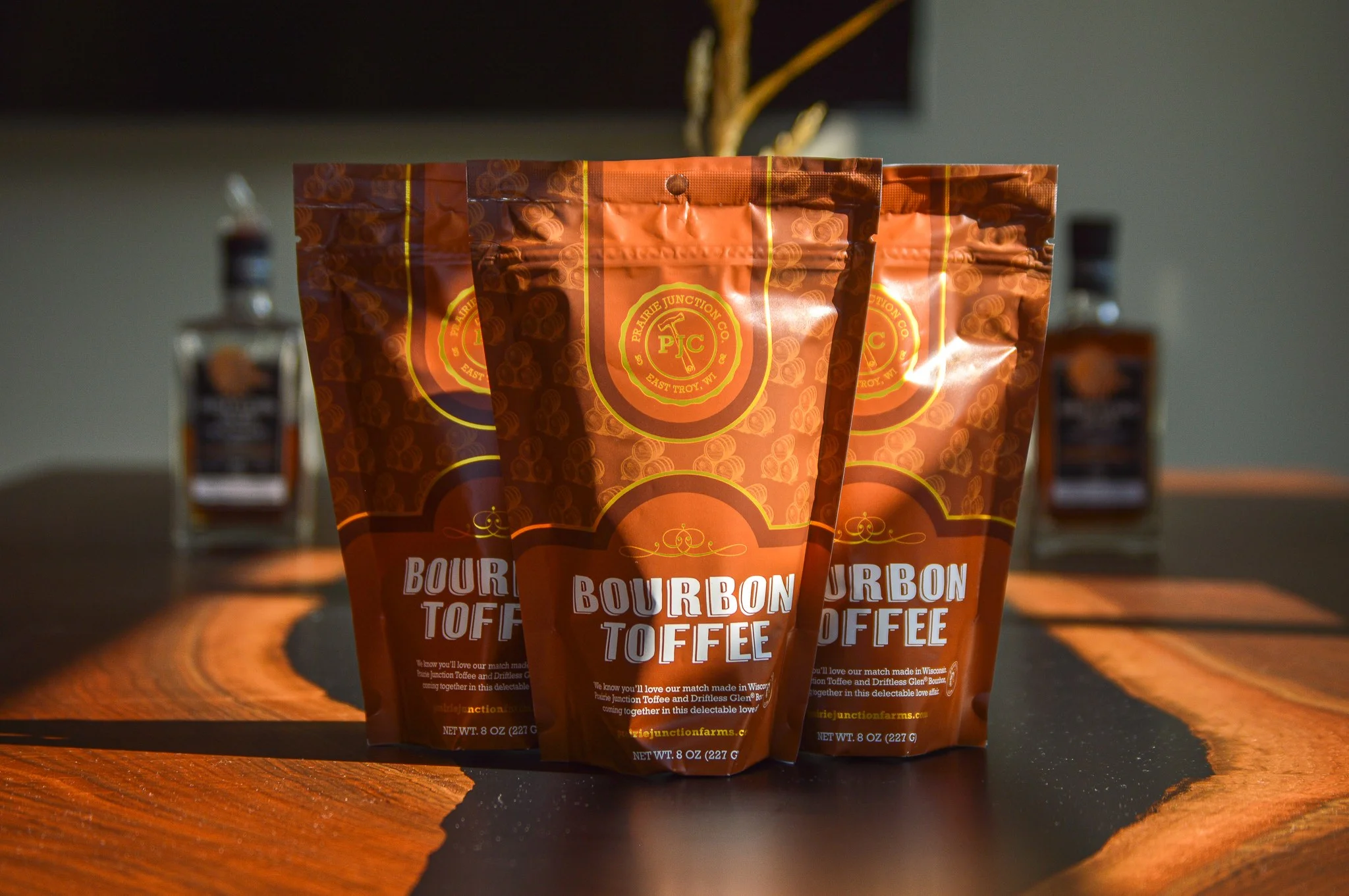 Three bags of bourbon toffee sit on a tabletop with bottles of bourbon in the background