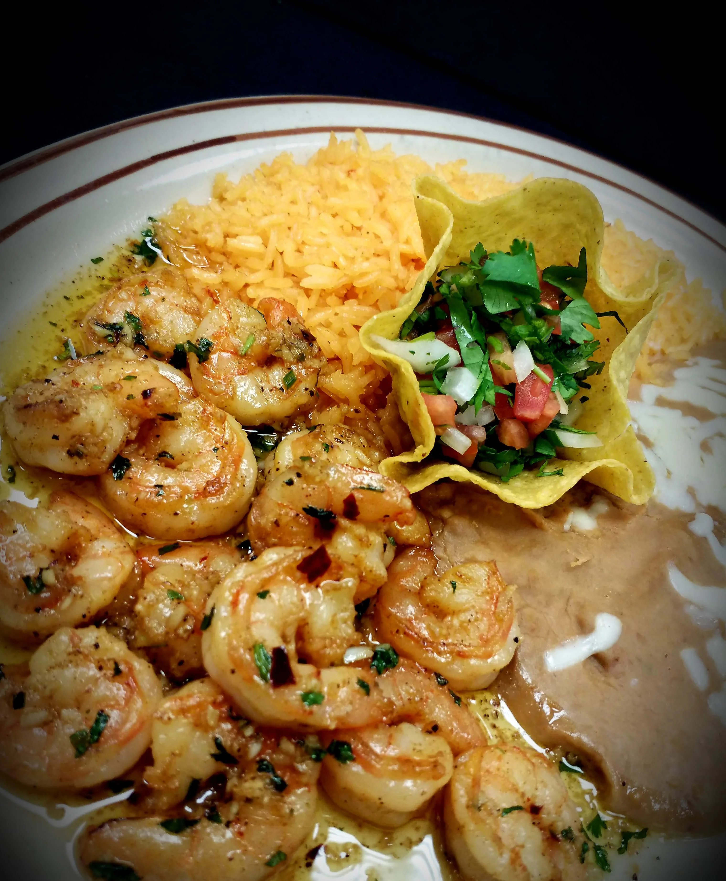 A plate of garlic shrimp served with Mexican rice, refried beans topped with cheese, and fresh pico de gallo.