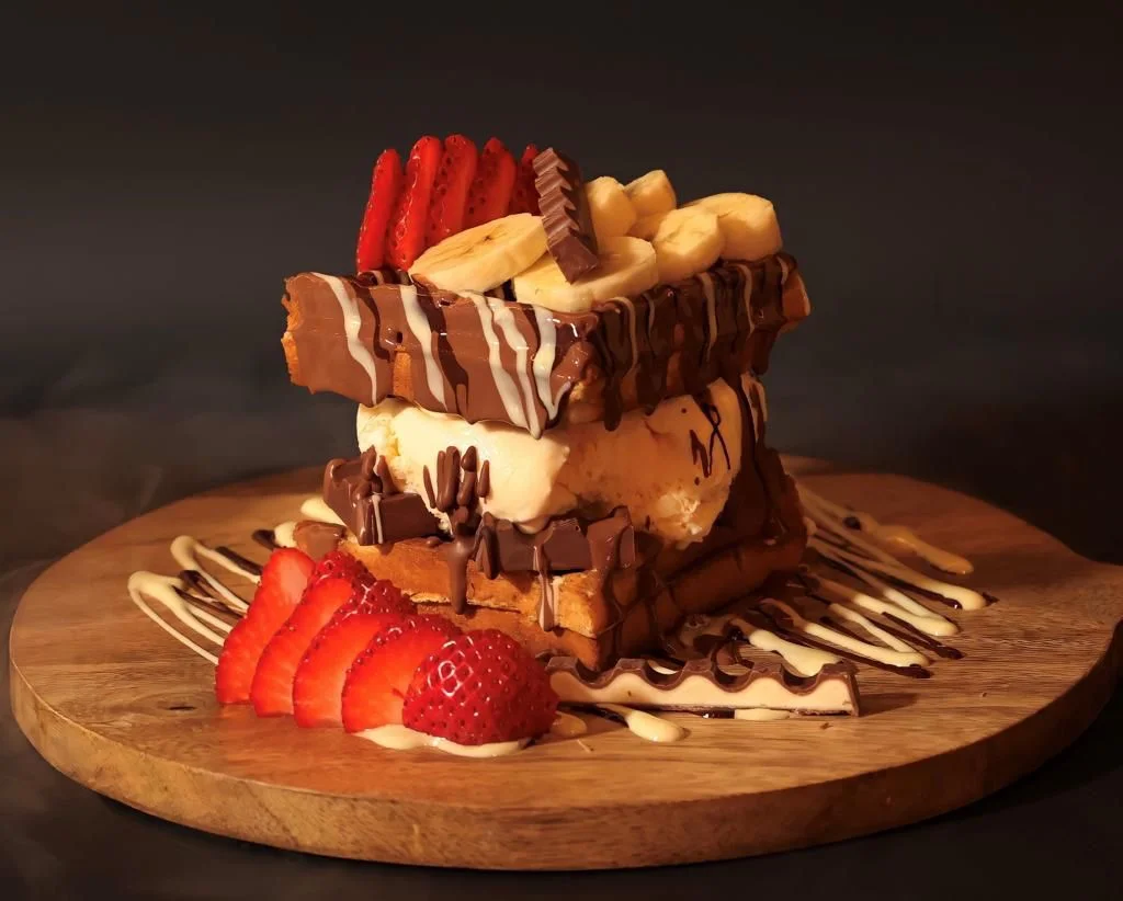 A triple-layered dessert with chocolate waffles, vanilla ice cream, fresh strawberries, and banana slices, beautifully arranged on a wooden platter.