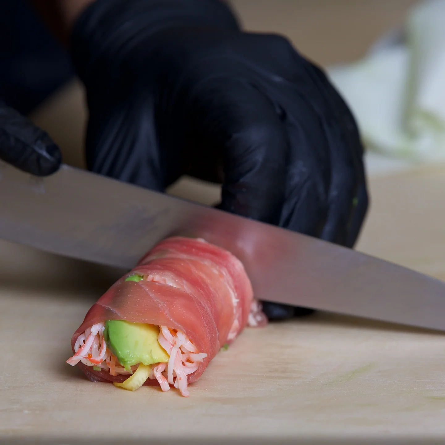Close-up of a chef in black gloves using a sharp knife to slice a sushi roll wrapped in fresh tuna and filled with avocado and crab.