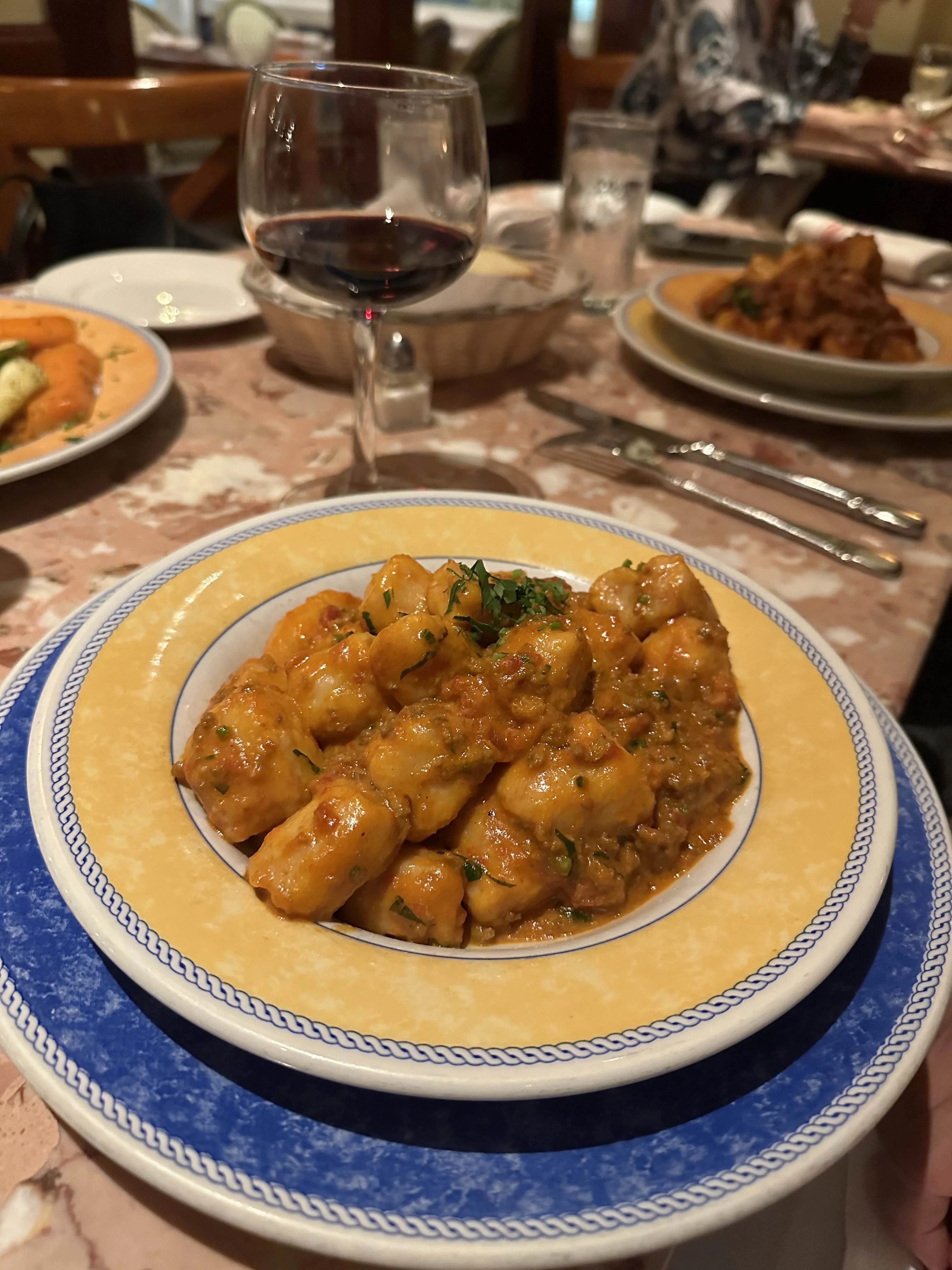A serving of gnocchi in a creamy tomato sauce garnished with herbs, presented in a decorative plate. A glass of red wine is in the background.