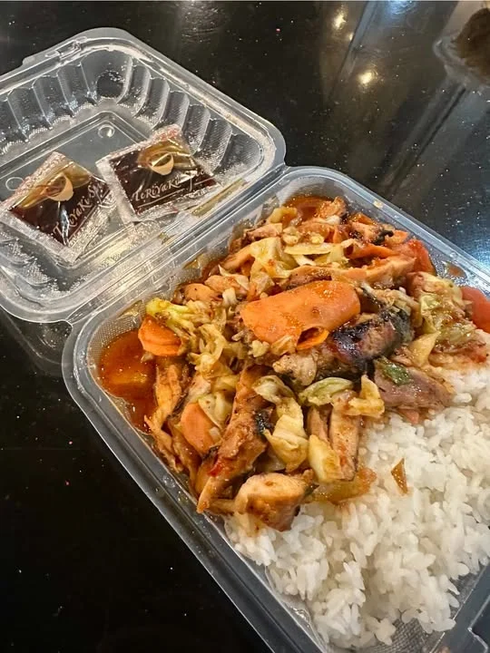 A meal box featuring rice and grilled chicken with vegetables, including cabbage and carrots, and two packets of sauce on top.
