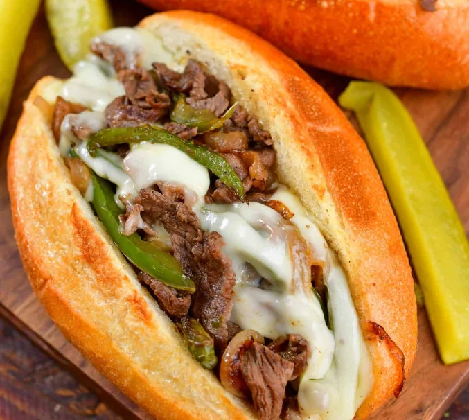 A delicious cheesesteak sandwich filled with sliced beef, green peppers, and melted cheese, served with pickles on the side.