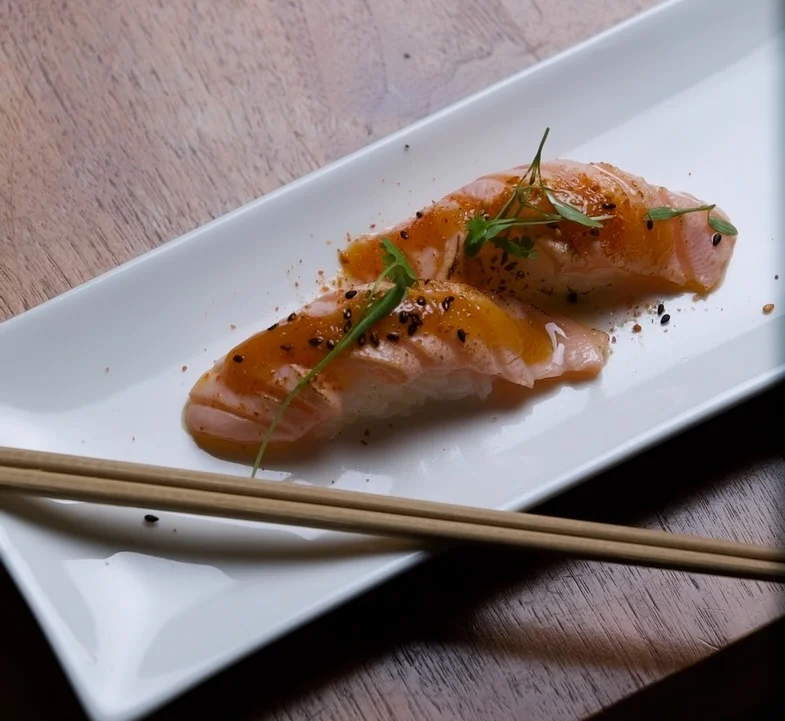 Two pieces of seared salmon nigiri sushi served on a long white rectangular plate, glazed with a sweet savory sauce, sprinkled with black sesame seeds, and garnished with microgreens, with a pair of chopsticks resting in the foreground on a wooden table.