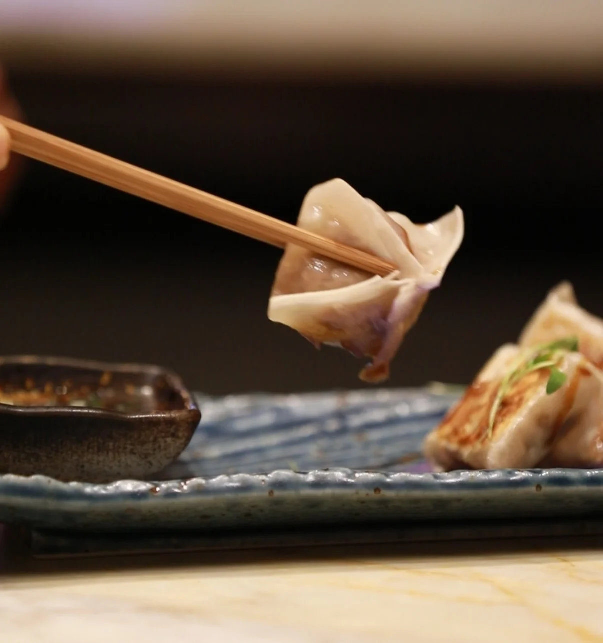 A single steamed or pan-fried dumpling being held up with wooden chopsticks over a textured blue plate containing more gyoza and a small dark bowl of dipping sauce.