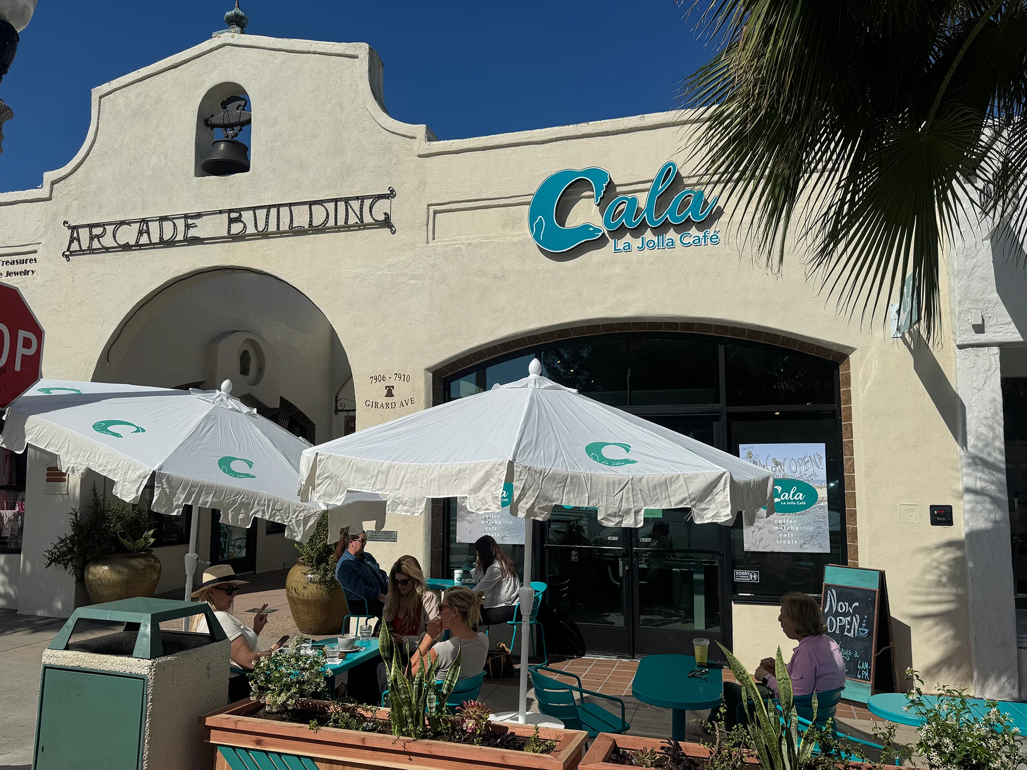 The exterior of a café with outdoor seating, featuring umbrellas and a vibrant sign, set against a clear blue sky.