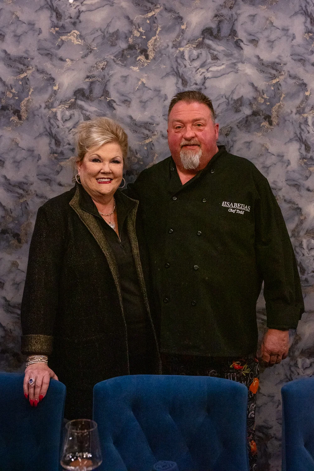 Chef Todd and Nickki Golden stand side by side smiling, both dressed in black chef coats. They are posing in front of a catering station with plated gourmet dishes, creating a warm and professional atmosphere.