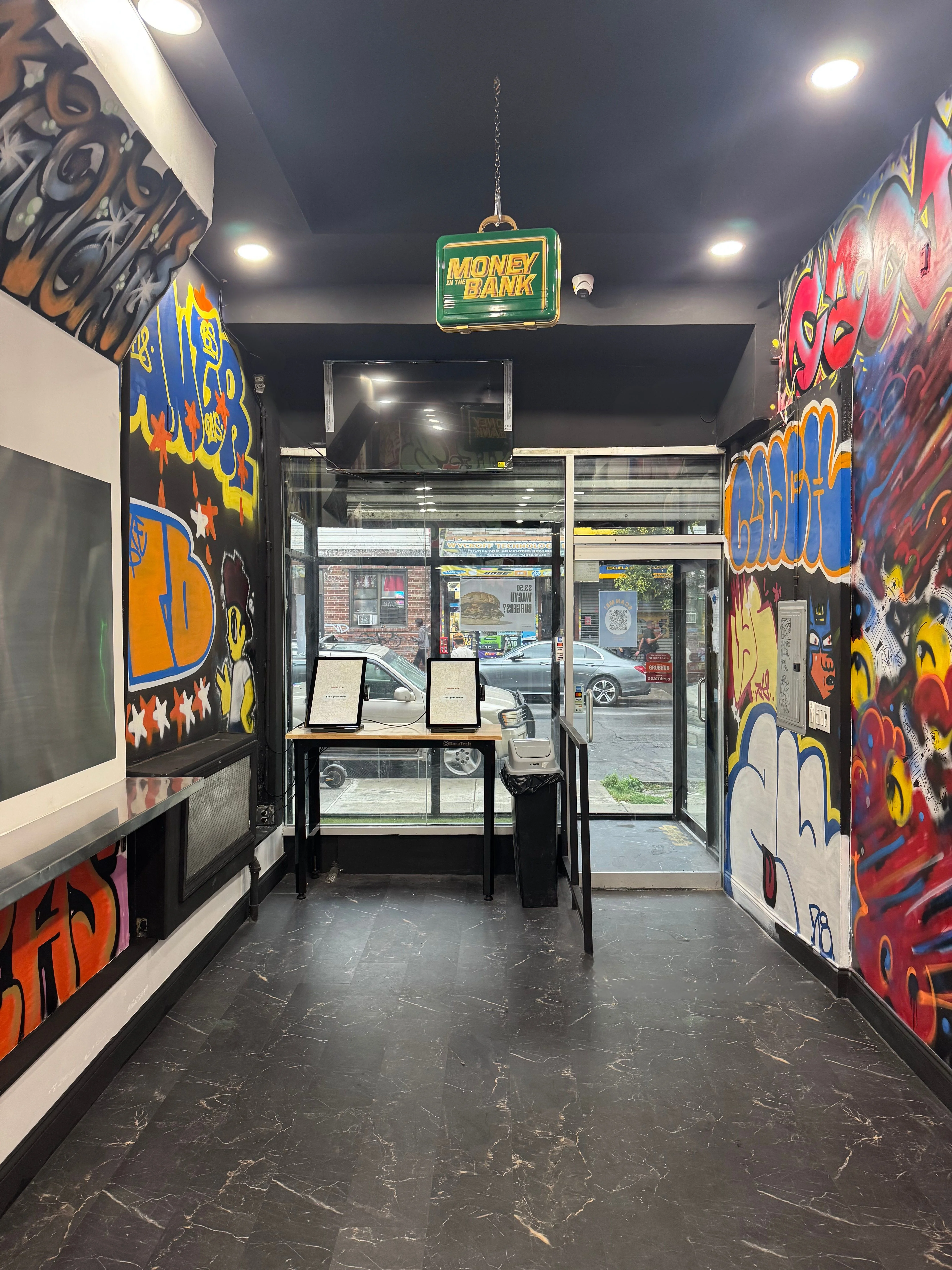 A lively space featuring graffiti art, a hanging sign labeled 'MONEY BANK', and a view of the street from large glass doors.