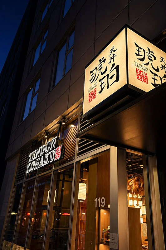 The exterior entrance of Tendon Kohaku at night, captured from a sharp upward angle. A brightly lit architectural cube sign projects from the building, casting a warm white radiance that contrasts with the night sky. The glass storefront below reflects subtle city lights while revealing a lantern-lit dining space inside.