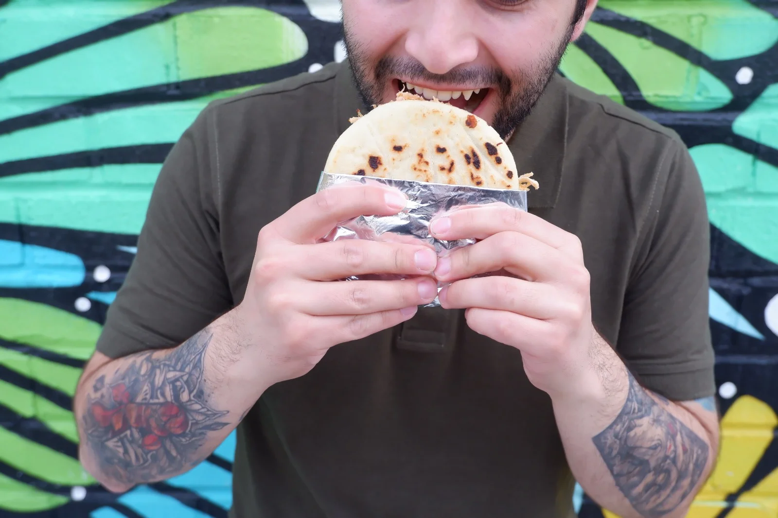 A person about to take a bite of a tortilla filled with ingredients, against a colorful mural background.