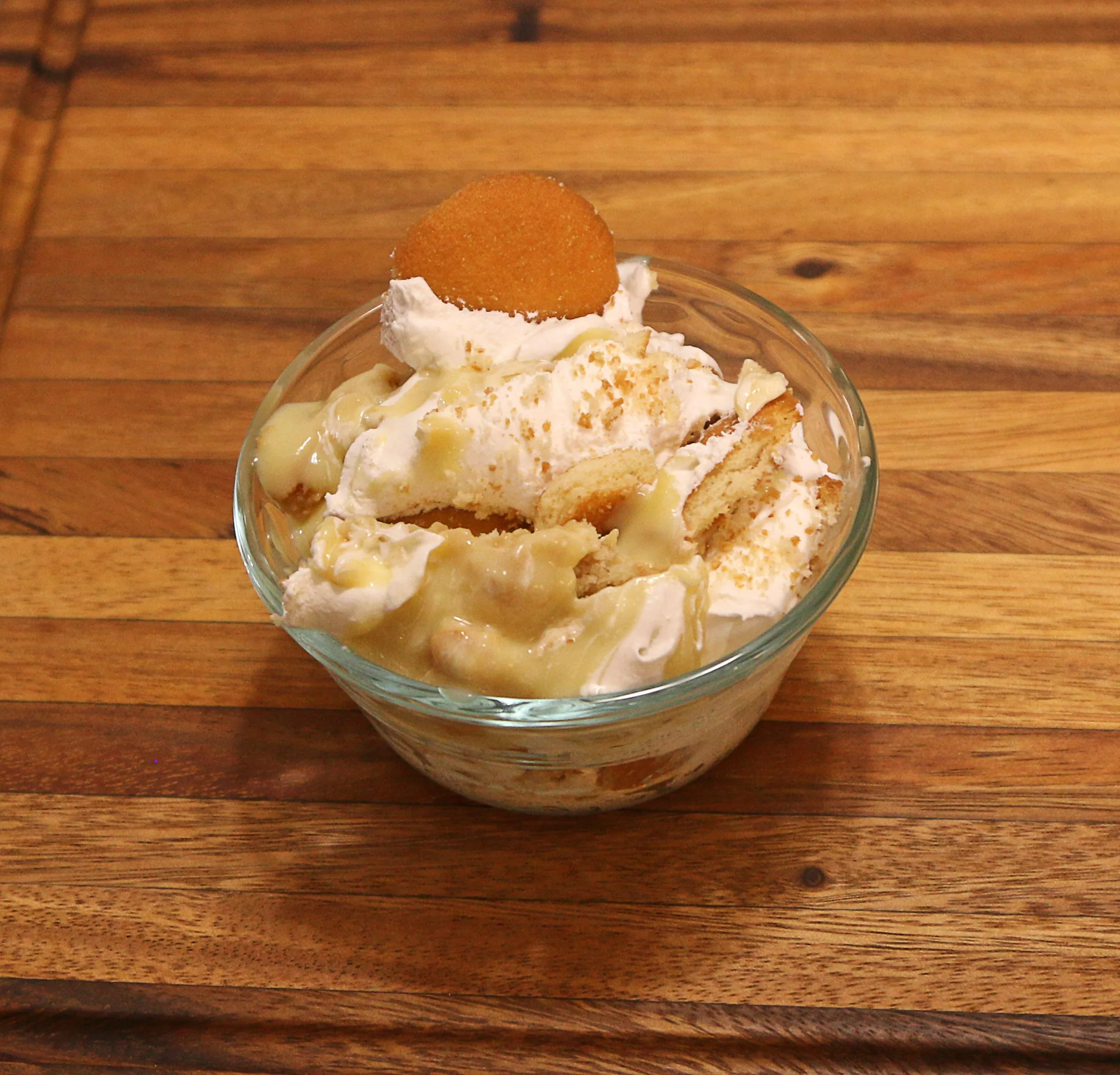 A glass bowl filled with a creamy dessert made of vanilla wafers, banana pudding, and whipped cream, topped with a wafer.