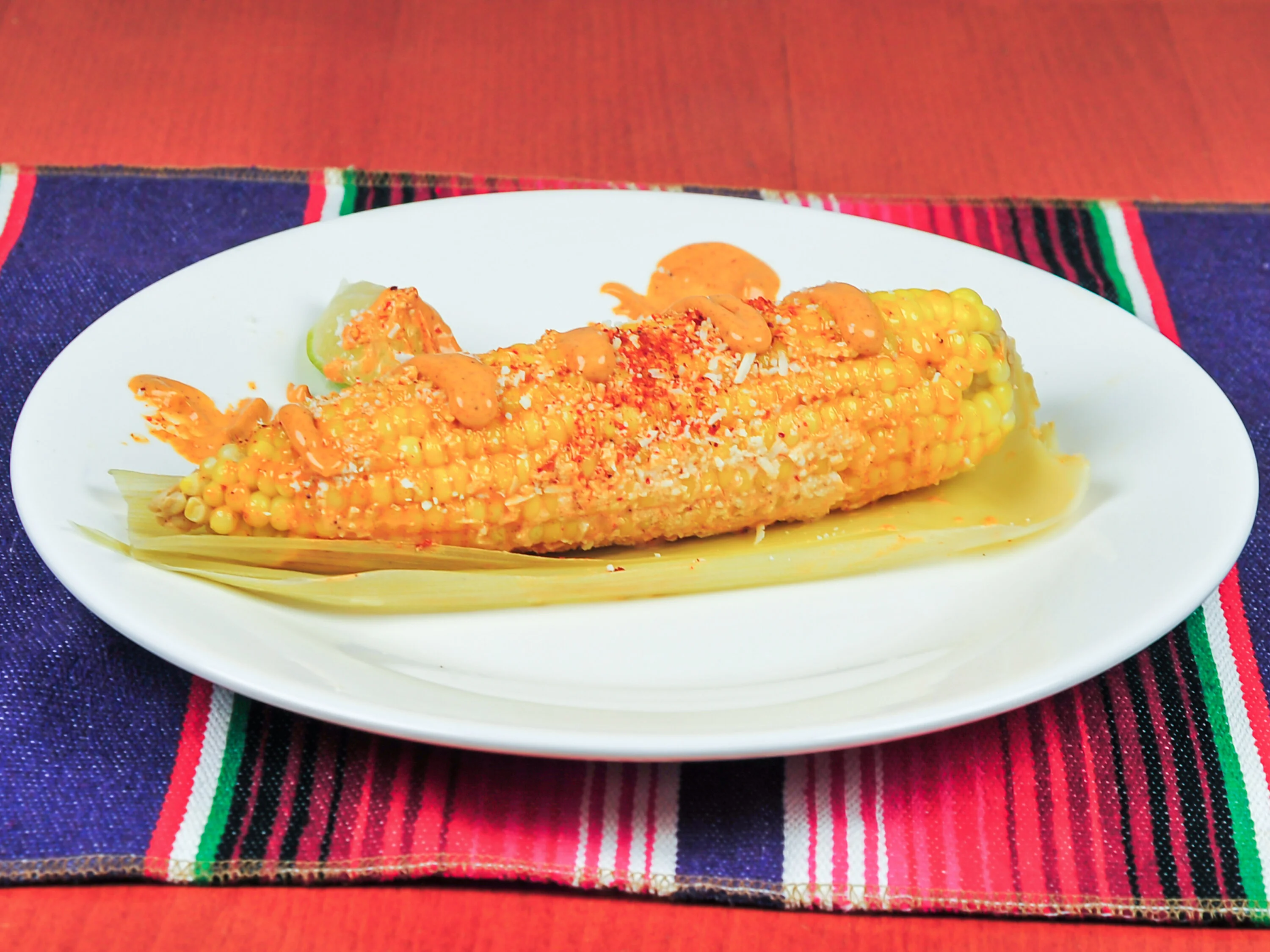 A plate of elote (Mexican street corn) topped with cheese, chili powder, and drizzled with sauce, served with a lime wedge.