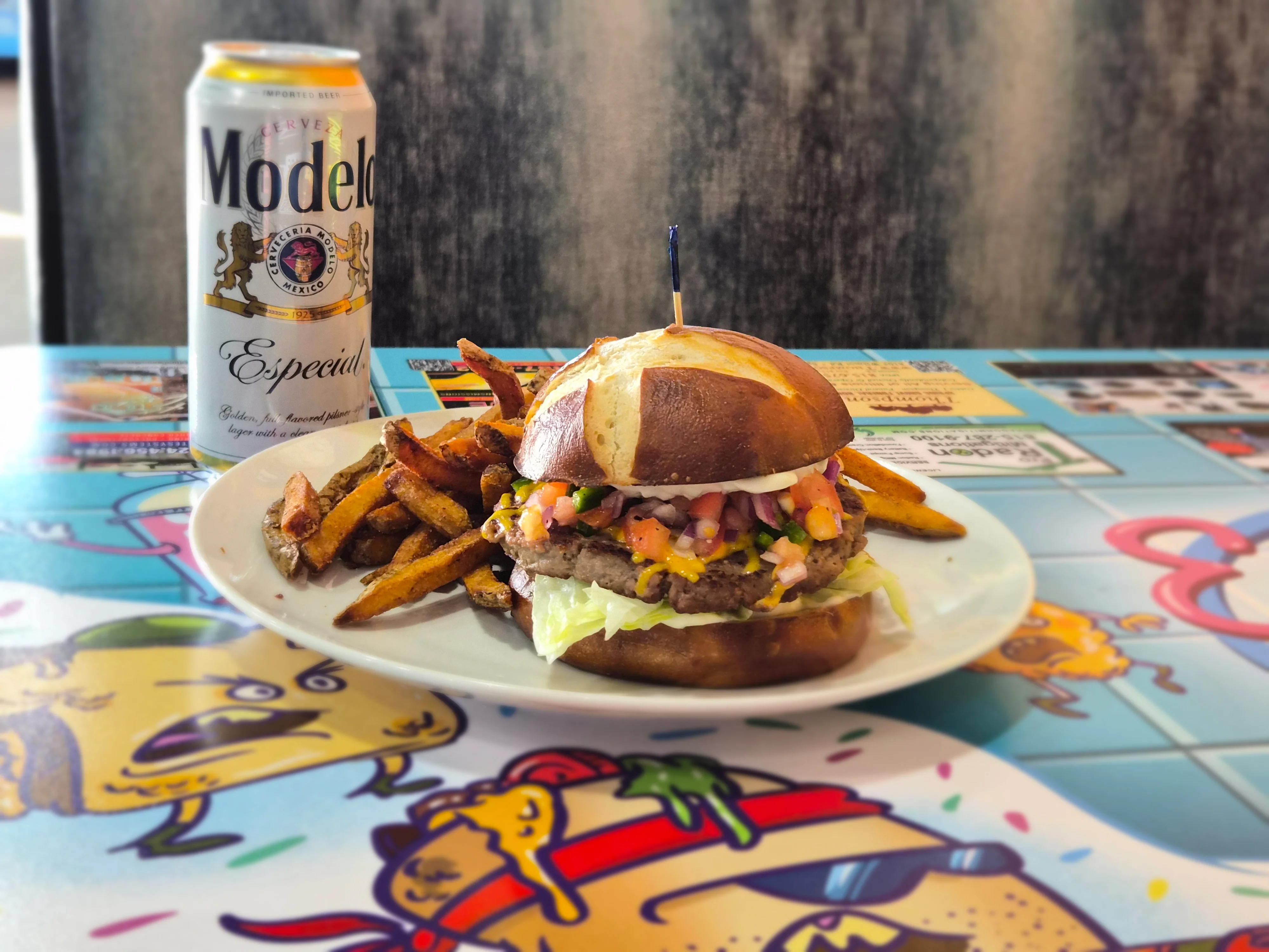 A delicious burger served with seasoned fries and a can of beer on a vibrant table cover.