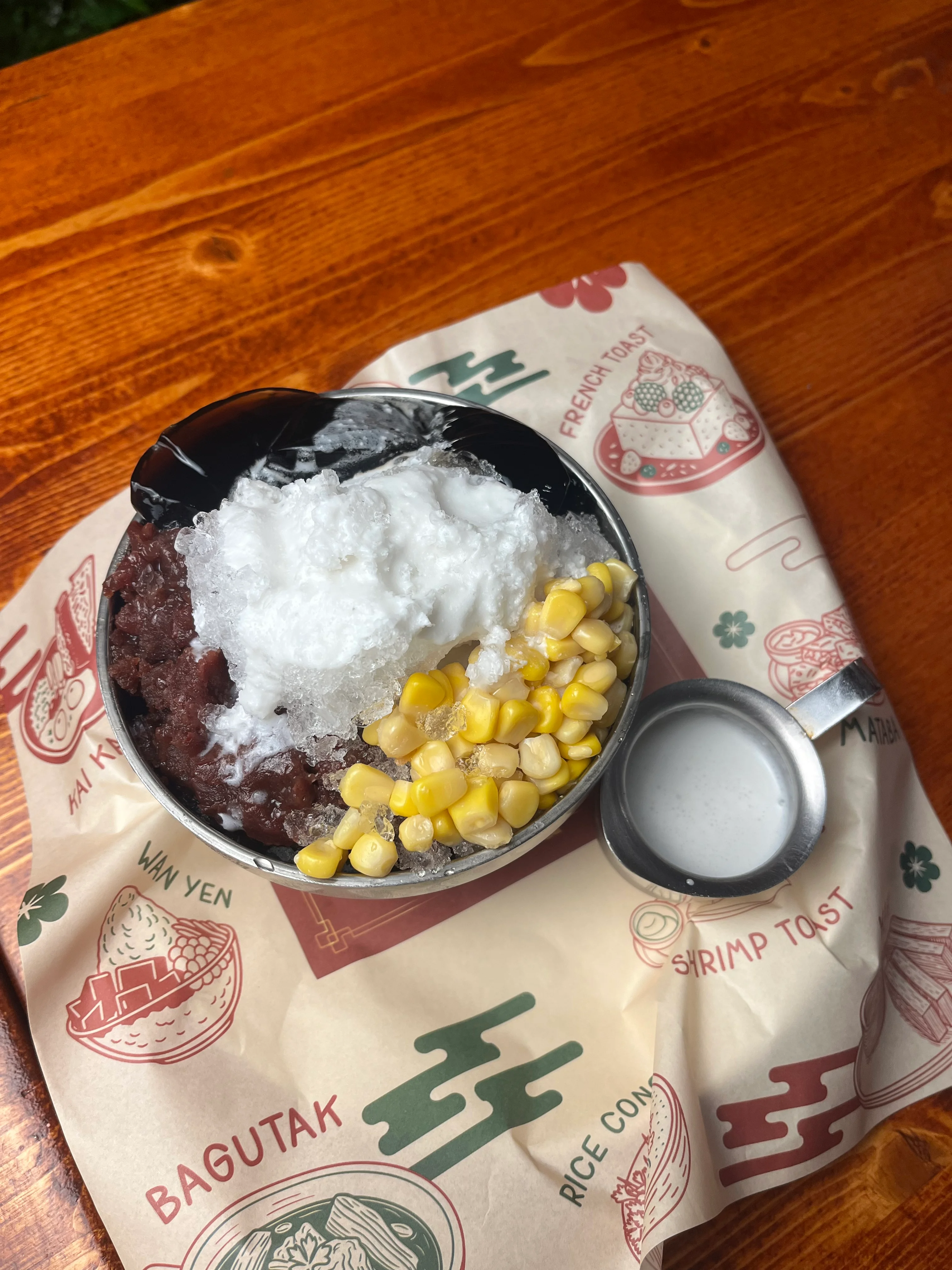 A bowl containing a mixture of sweet red beans, shaved ice, sweet corn, and topped with a generous amount of coconut cream, served on a patterned paper.