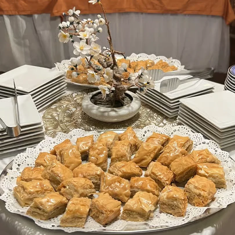 A catering display of golden-brown, honey-glazed Baklava pieces arranged on a silver platter with white lace doilies, featuring a decorative floral centerpiece and stacked plates.