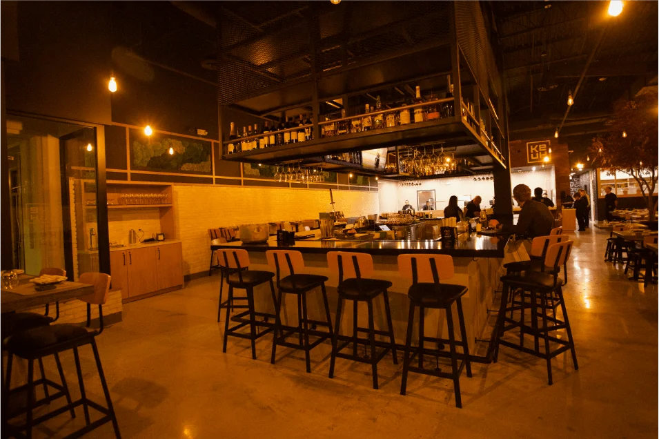 Boca Raton bar dimly lit featuring warm hanging light bulbs, modern wooden bar stools lined along a sleek dark counter, and a large black metal overhead rack stocked with wine and liquor bottles.