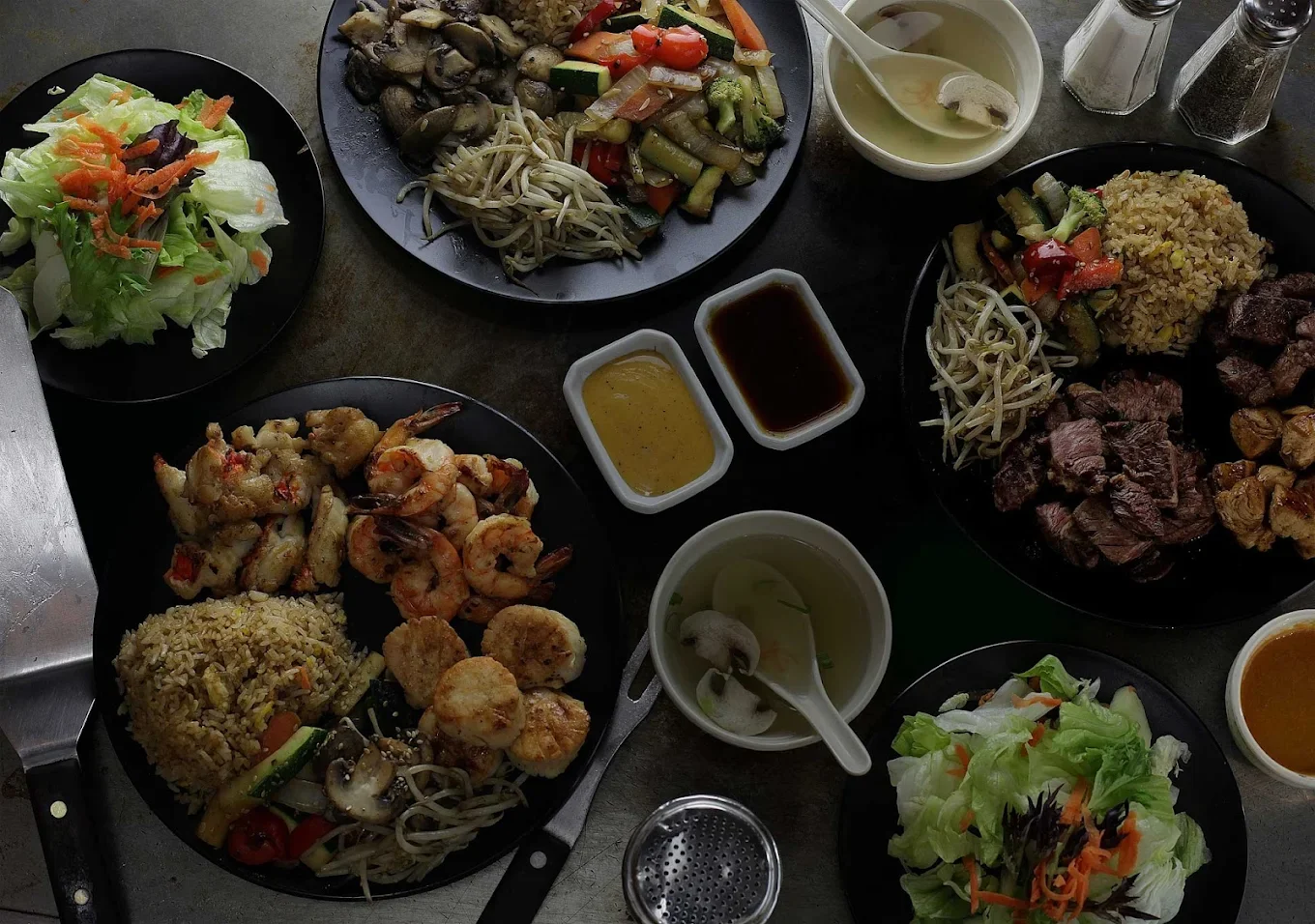 An assortment of Asian dishes on black plates, including fried rice, vegetables, shrimp, and sauces, arranged for a meal.