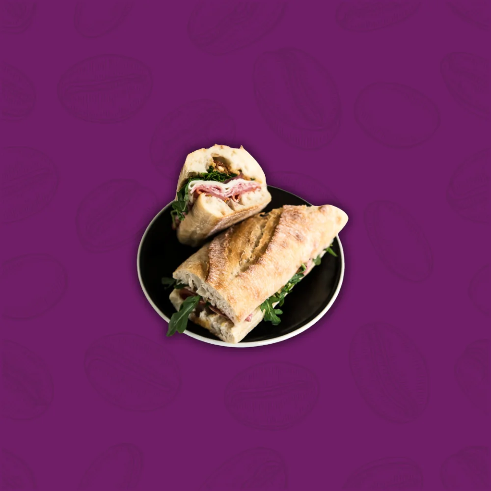 A sandwich with deli meat and greens on ciabatta bread is halved on a black plate, set against a purple background with subtle patterns.