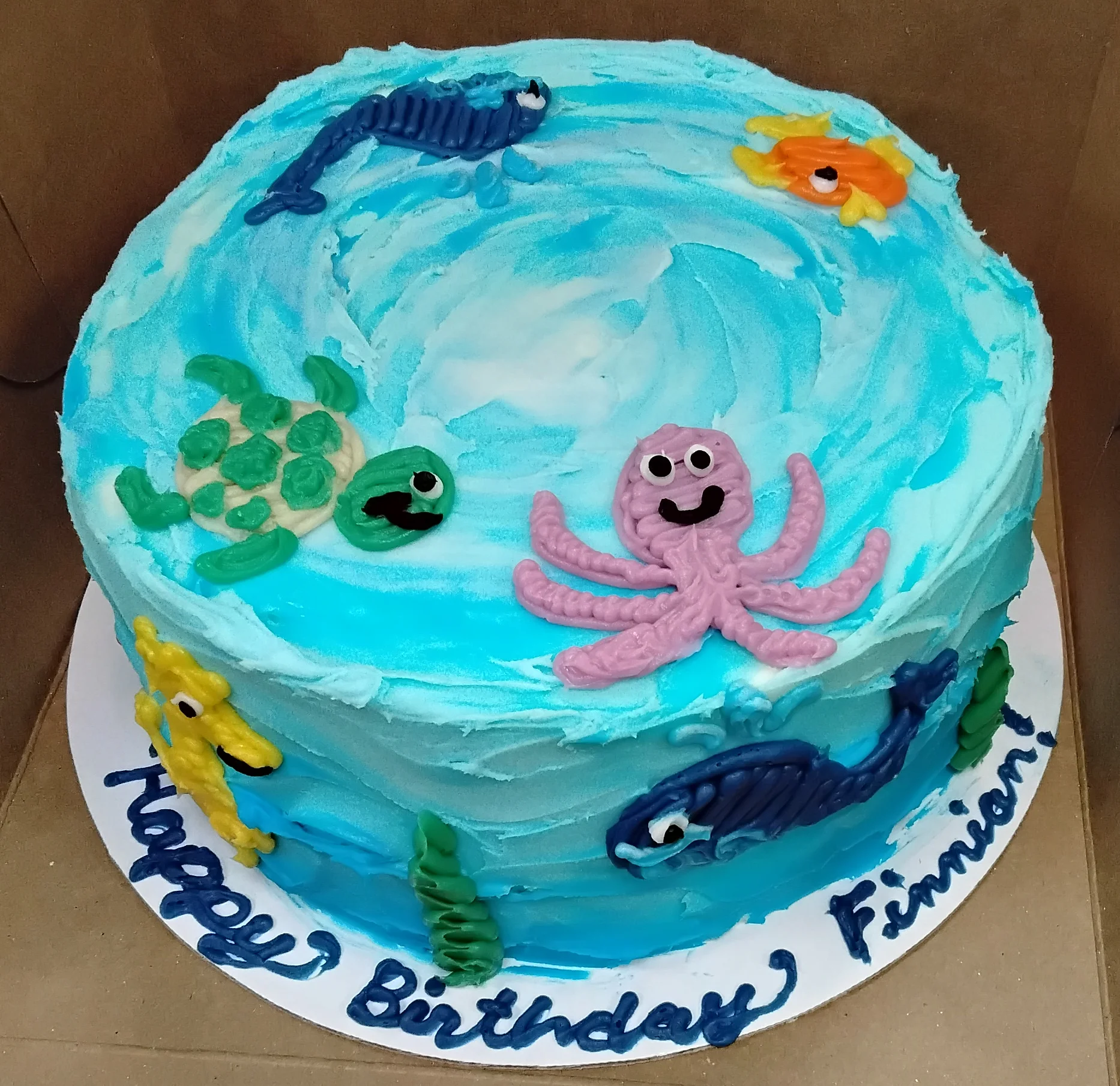 Round cake with ombre icing to look like an ocean with several sea creatures drawn in icing and text that says "Happy Birthday Finnian!"