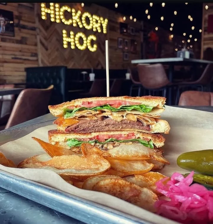 A hearty sandwich stacked with meat, lettuce, and tomato, served alongside crispy potato chips and pickles. The restaurant ambiance features warm lighting and rustic decor.