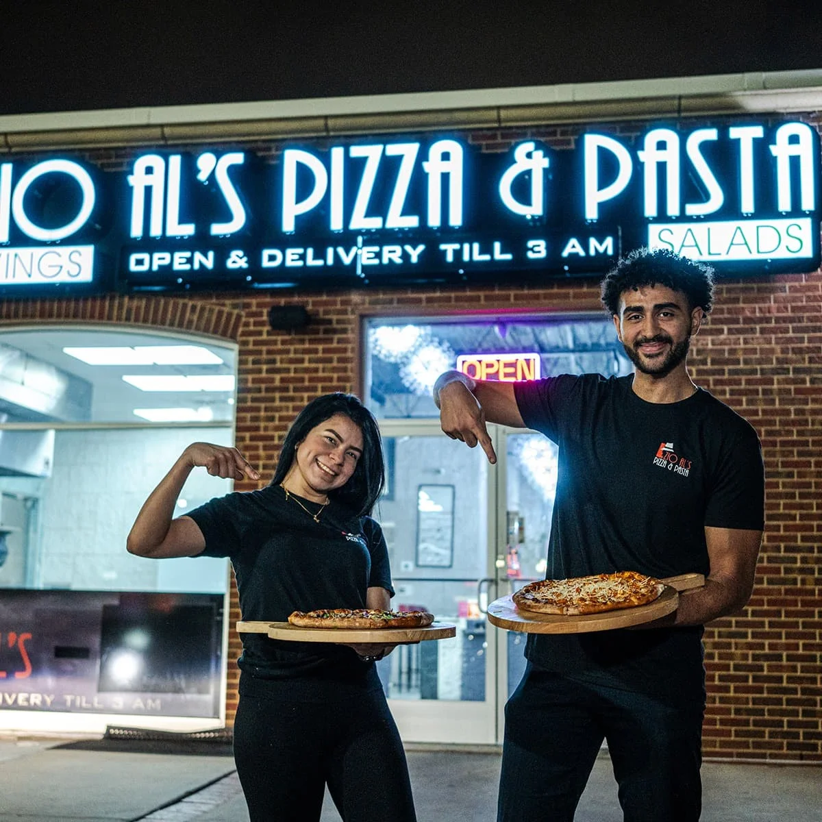 Two staff members showcasing pizzas outside a lively pizzeria
