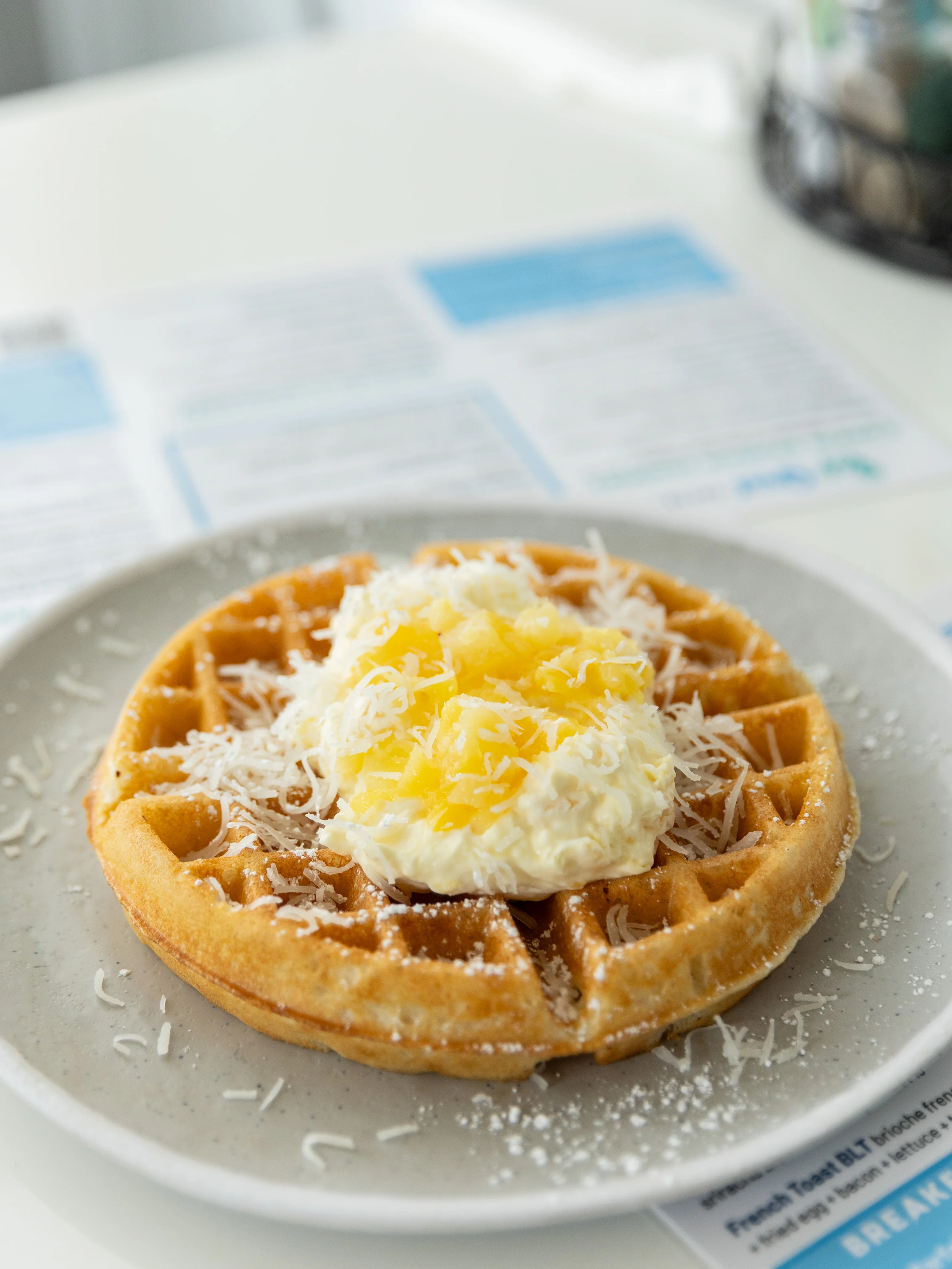 A golden-brown waffle topped with whipped cream, pineapple, and shredded coconut, served on a simple plate.