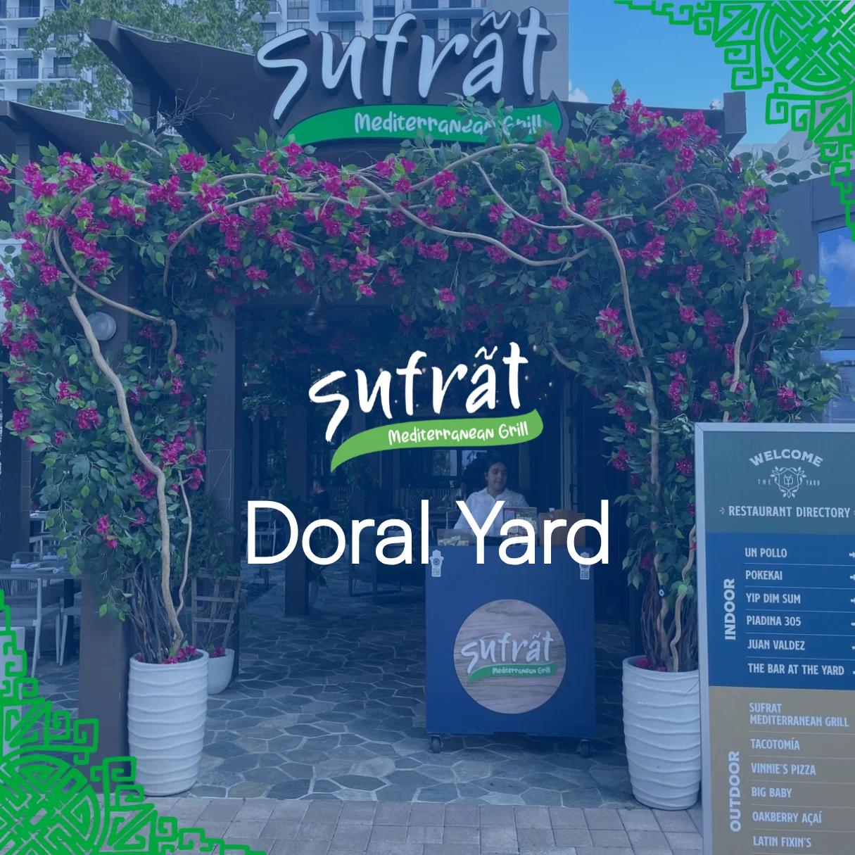 Entrance to Sufrat Mediterranean Grill at Doral Yard, adorned with vibrant pink flowers. The sign shows the restaurant directory beside it.