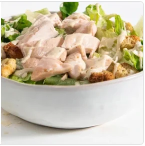 A bowl of Caesar salad topped with sliced chicken and croutons, drizzled with dressing.