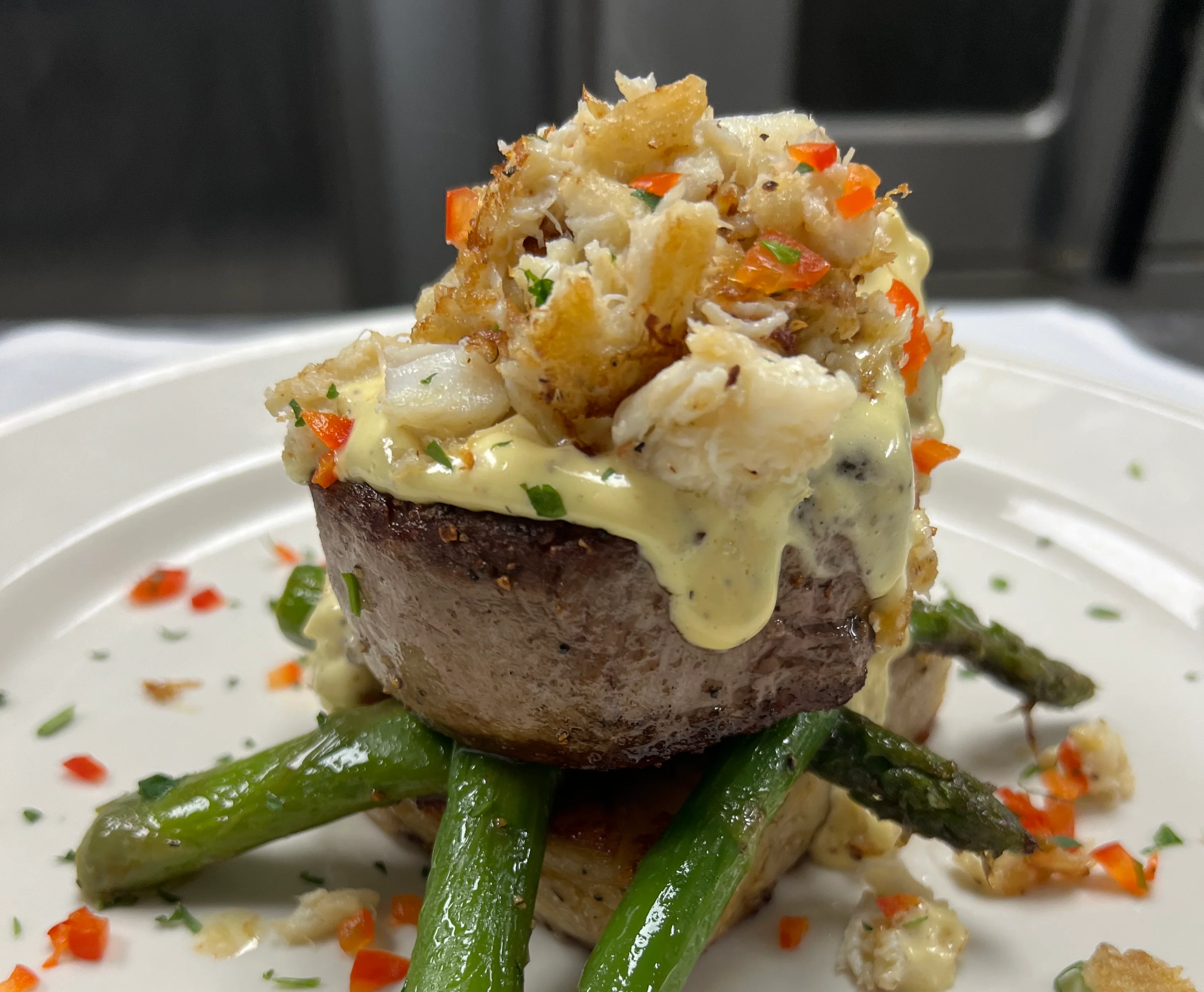 A gourmet entrée featuring a filet mignon topped with lump crab meat and a creamy hollandaise-style sauce, served on a bed of seared asparagus spears. The dish is garnished with finely chopped red peppers and herbs on a white plate, with a drizzle of sauce and scattered crab pieces enhancing the elegant presentation.