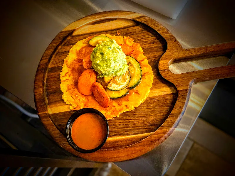 A colorful dish featuring a golden tortilla topped with sliced zucchini, orange slices, and a generous dollop of guacamole. Accompanied by a small bowl of sauce on a wooden serving board.