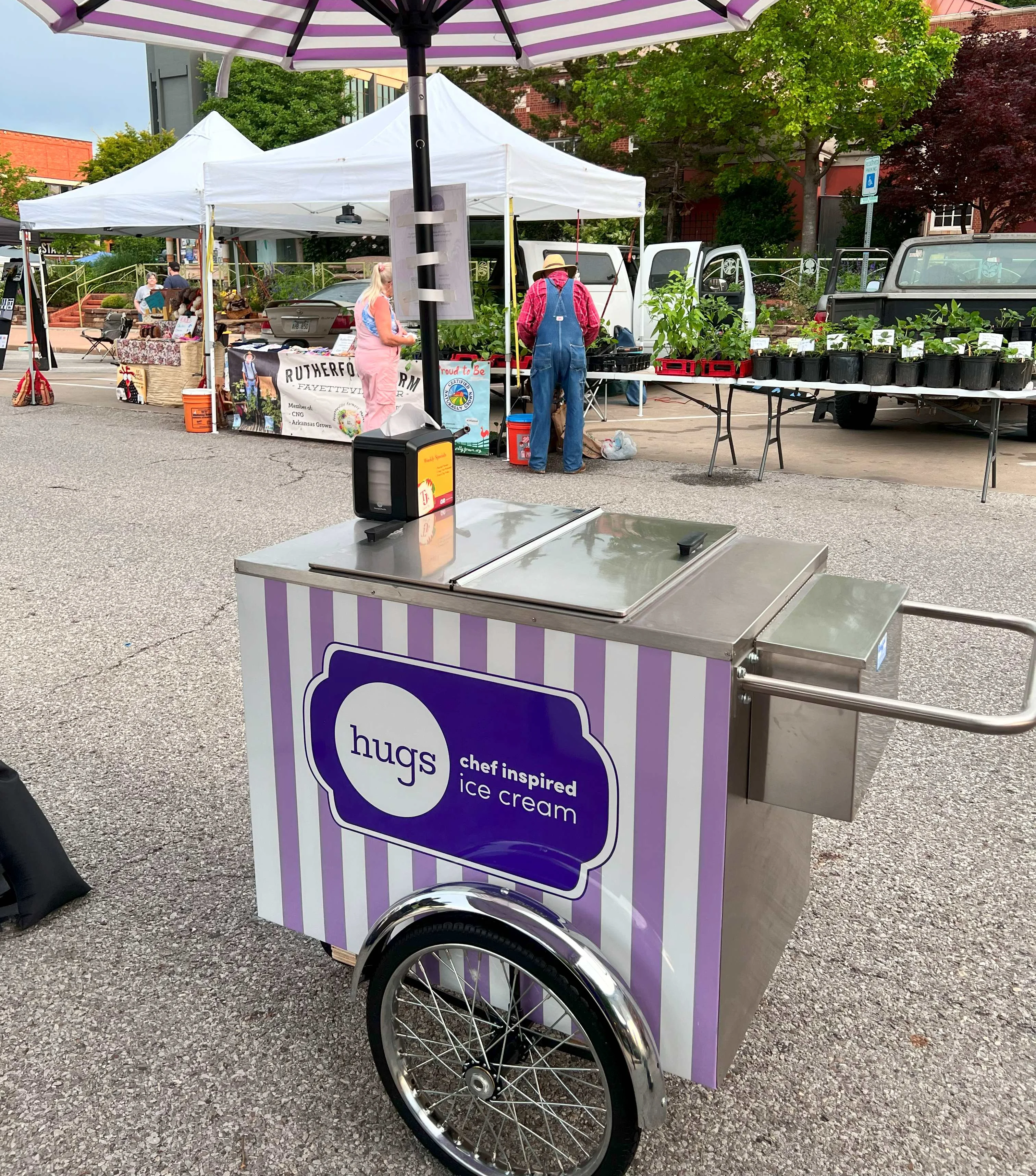 Hugs Ice Cream mobile cart with purple and white stripes at a local farmers market, offering chef-inspired ice cream under a matching umbrella surrounded by vendor tents and shoppers