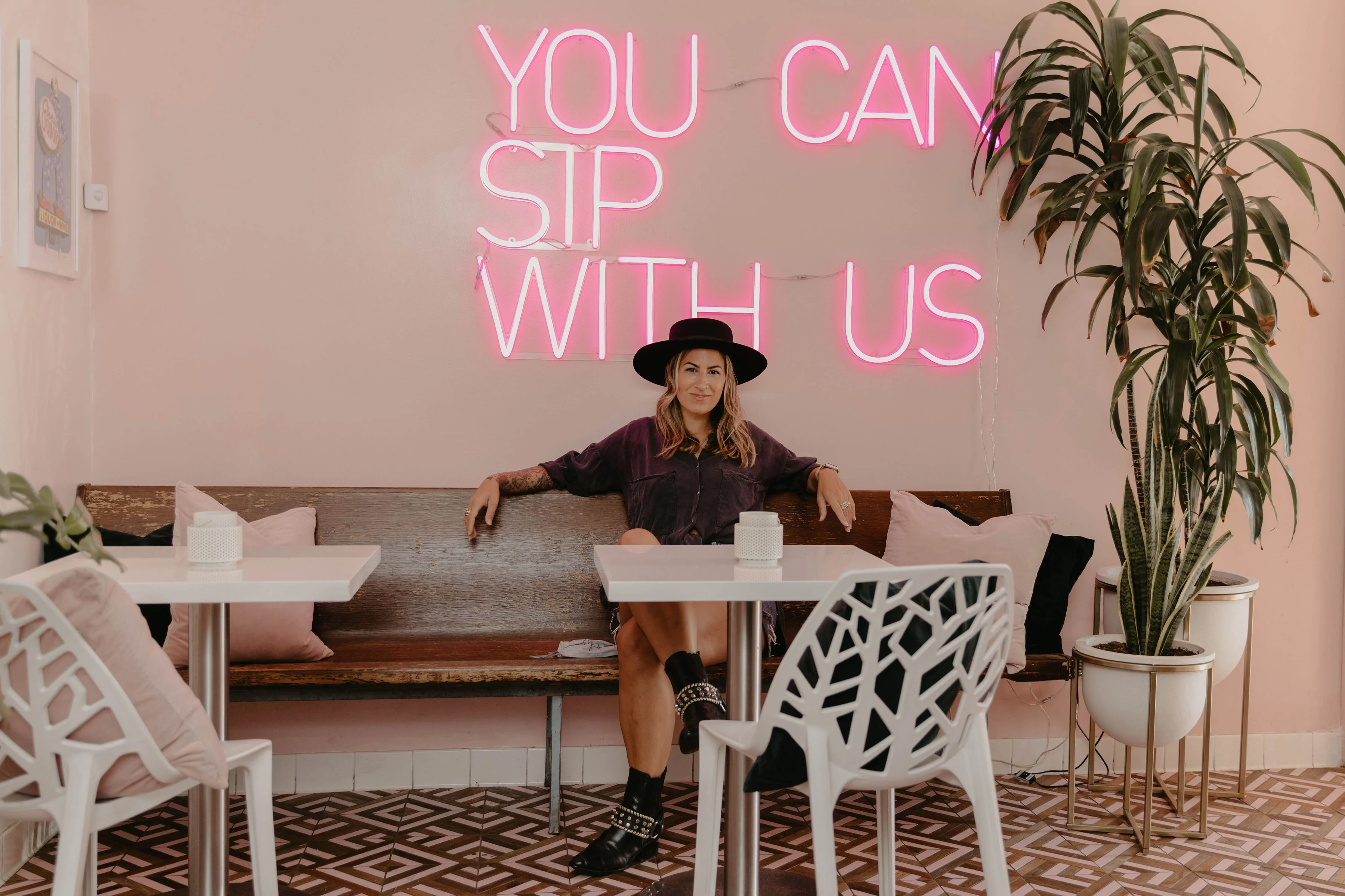 Girl sitting at a table with "you can sip with us" neon sign above her