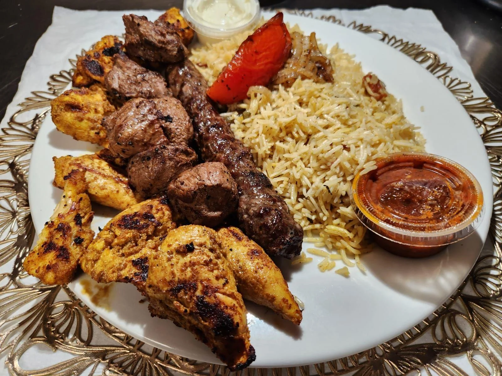 A mixed grill platter with chicken and beef kebabs, served with rice, a grilled tomato, and dipping sauces.