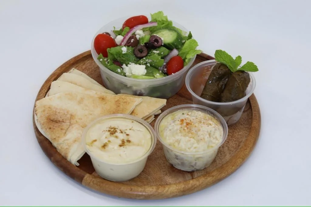 A wooden plate featuring a variety of Mediterranean dishes, including pita bread, a fresh salad with vegetables, and small containers of dips and stuffed grape leaves.