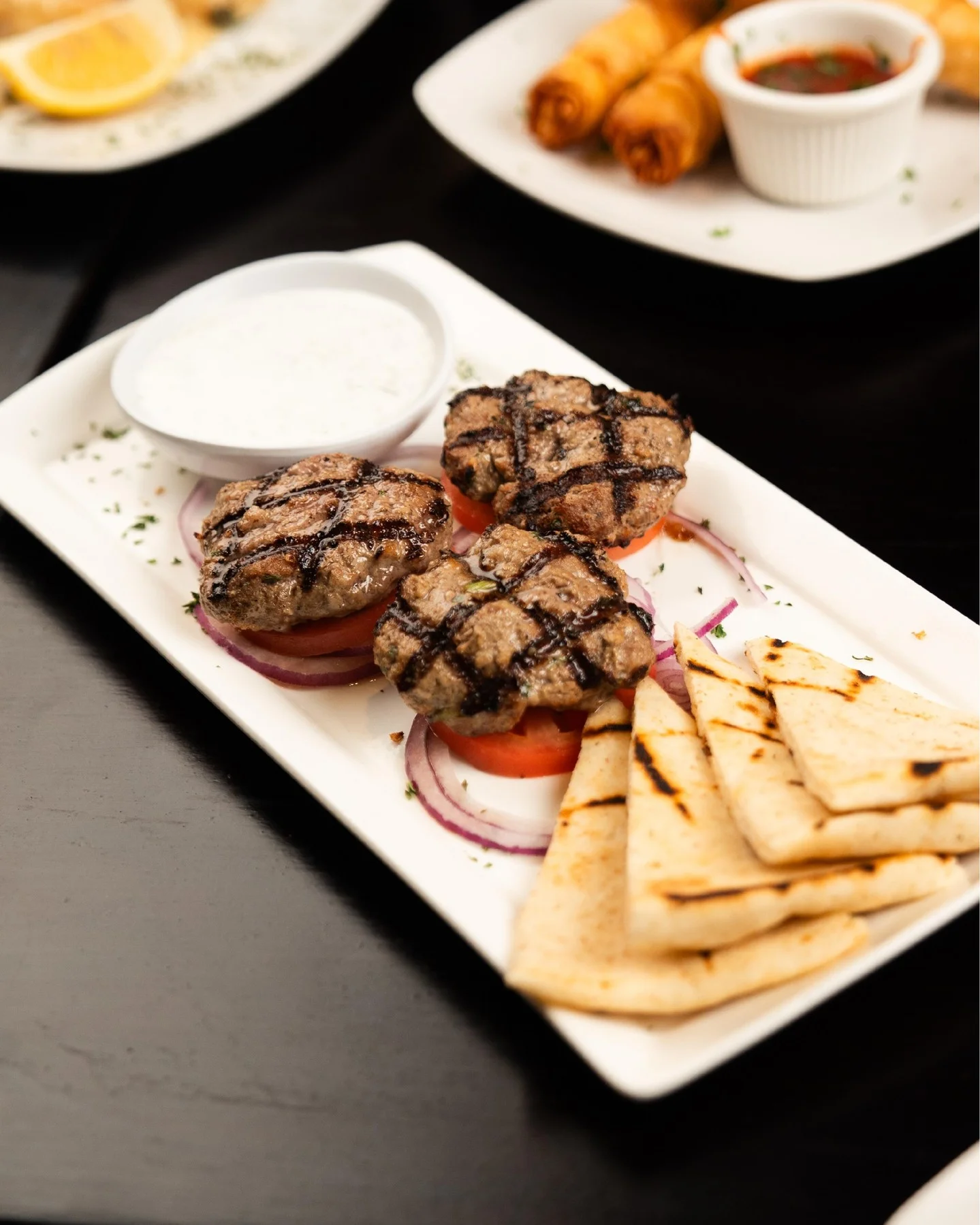 A serving of grilled meat patties accompanied by fresh vegetables and bread, arranged on a rectangular plate, with sauces in the background.