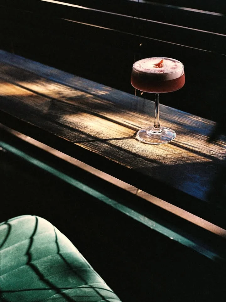 A cocktail glass on a wooden surface, illuminated by natural light, showcasing a vibrant drink topped with foam and a garnish.