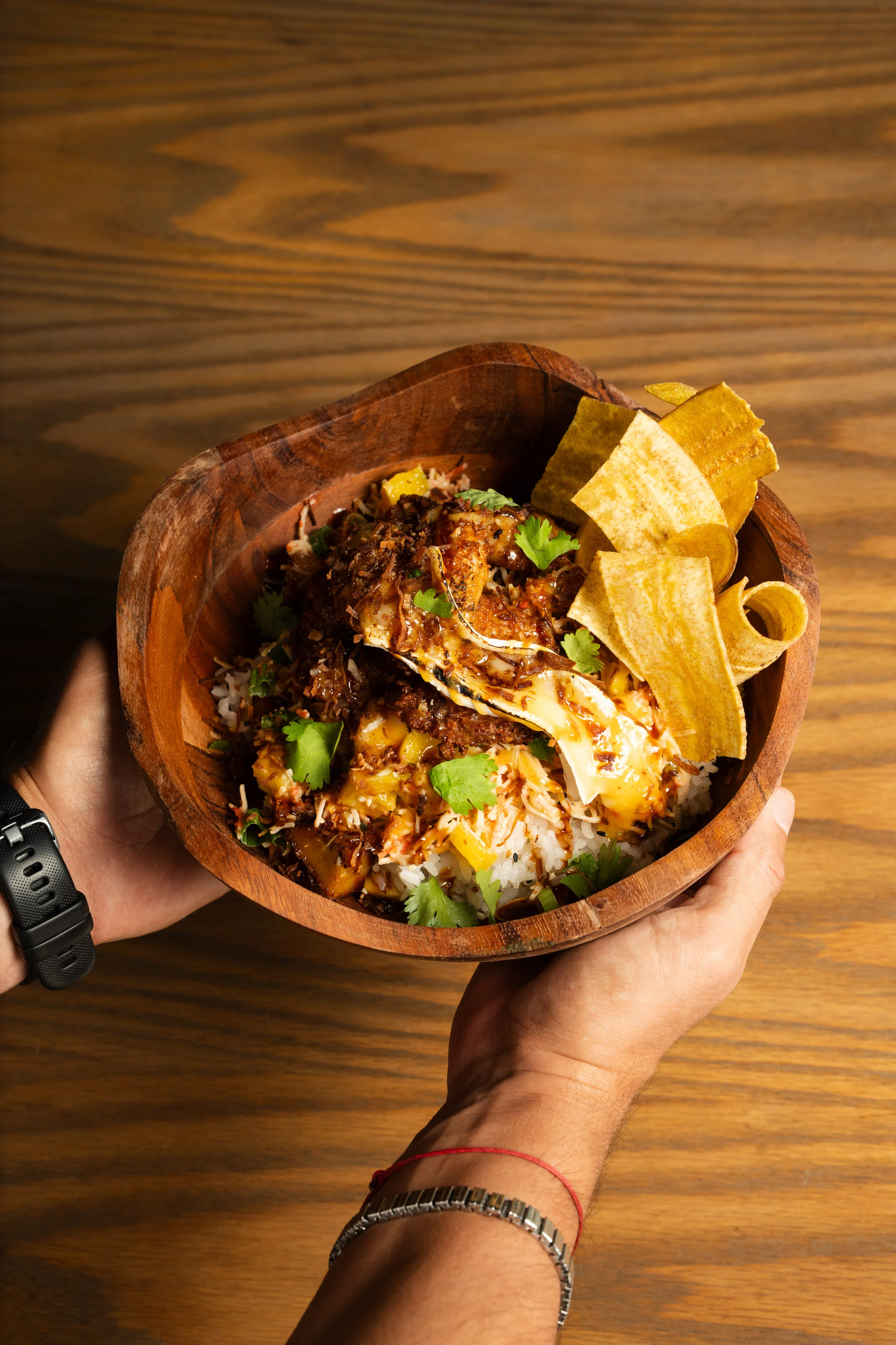 A wooden bowl filled with rice, topped with shredded meat, fresh herbs, and plantain chips, held by a hand.