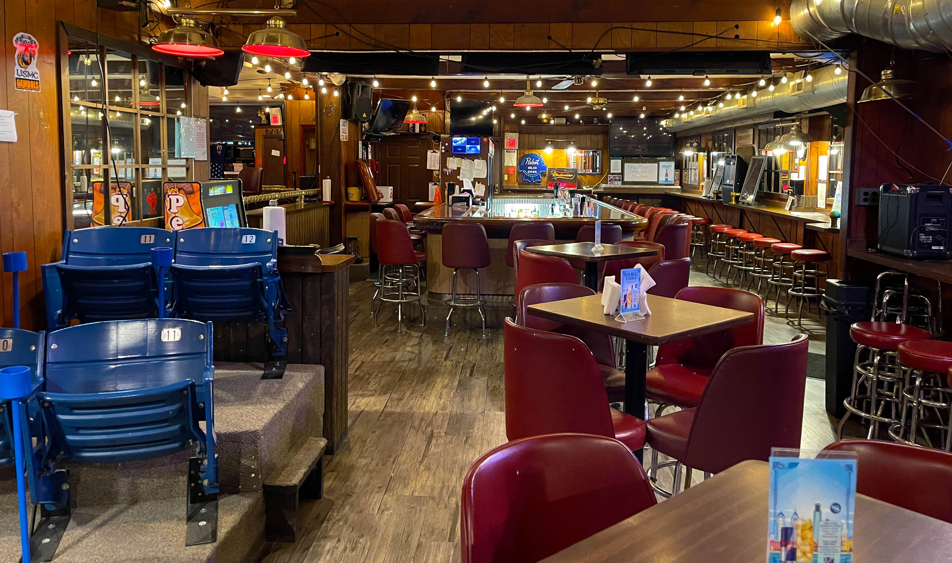 An inviting bar area with wooden decor, featuring red seating and a well-lit bar. The setting has a cozy ambiance with string lights above.