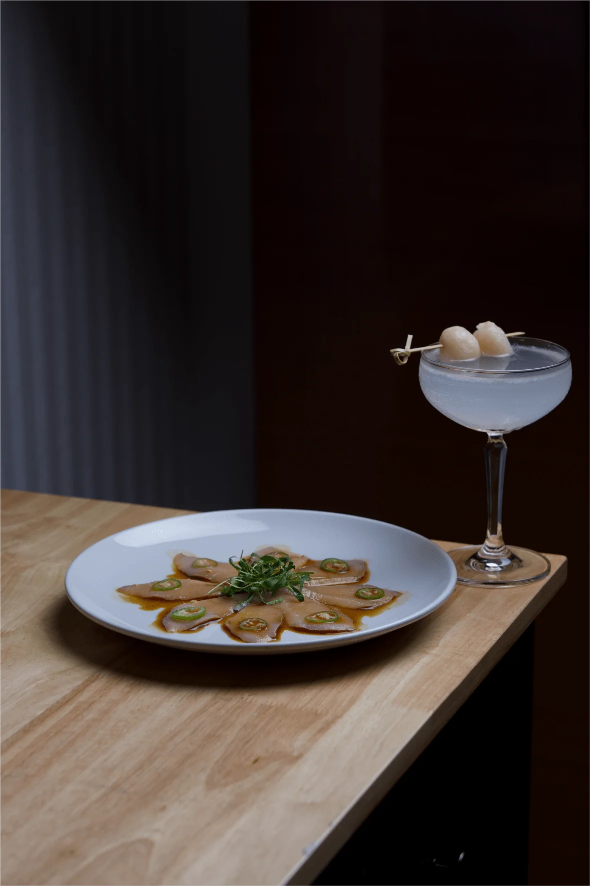 A sushi roll sliced into eight pieces, filled with fish and avocado, topped with thin slices of jalapeño and microgreens, and served on a long rectangular white plate with a dark sauce drizzle, resting on a table with a branded placemat.