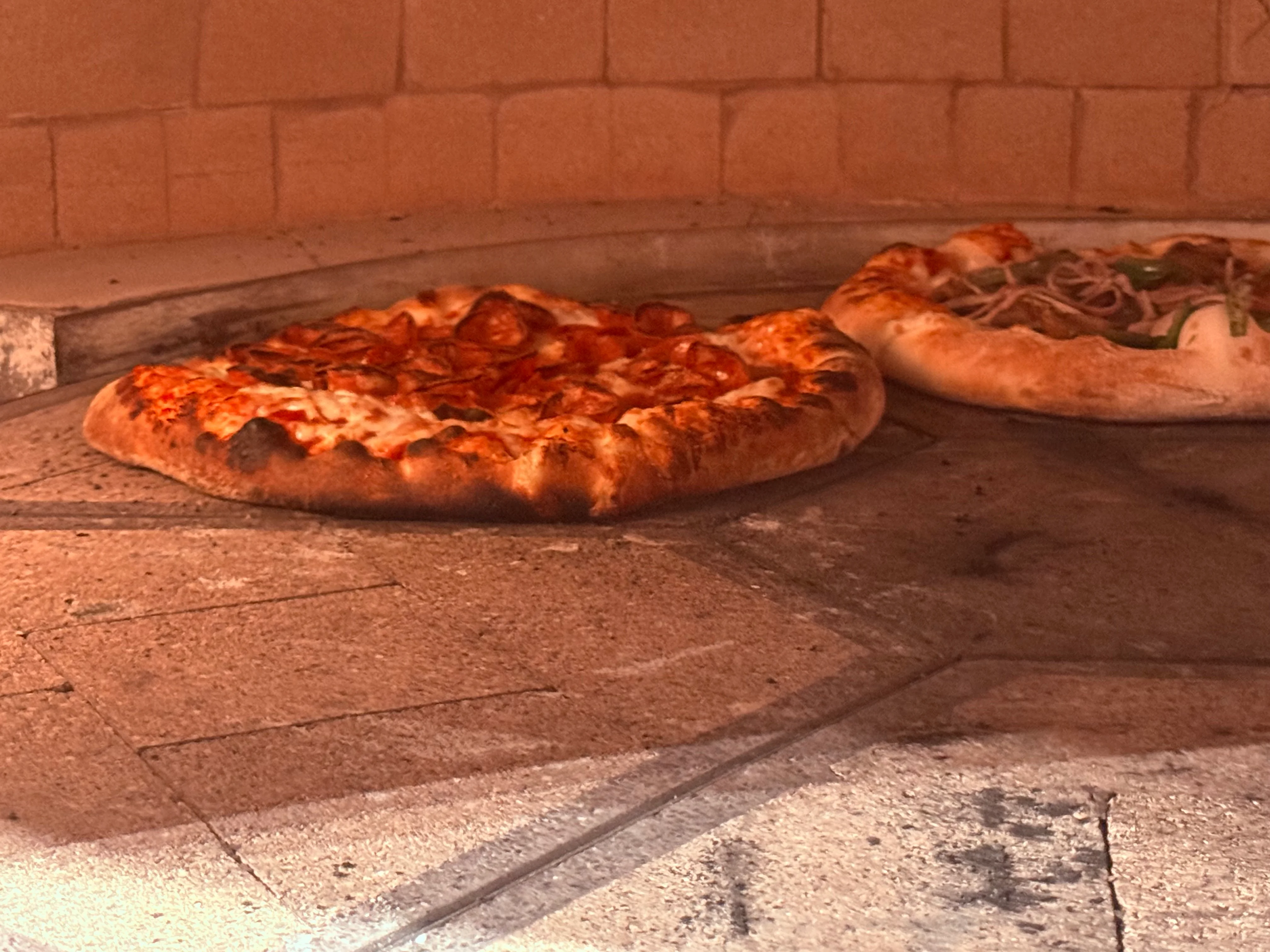 A chef holding a freshly baked pizza with a golden crust and green toppings in front of a pizza oven.