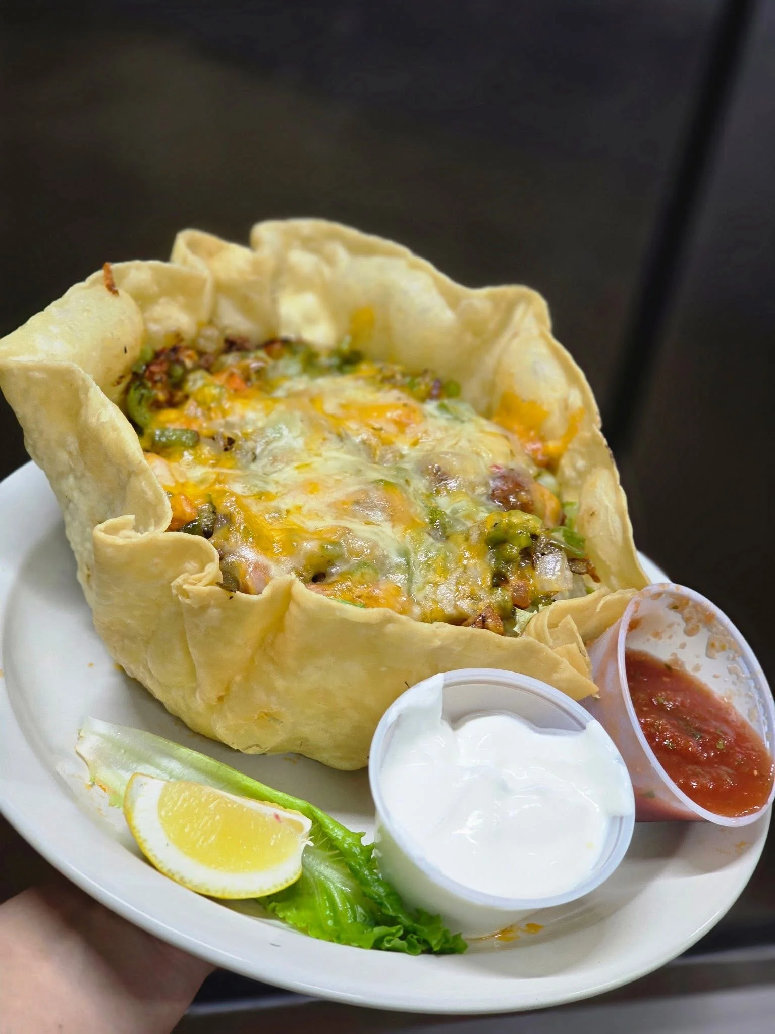 A baked tortilla bowl filled with a variety of ingredients including cheese, vegetables, and meat, served with sides of sour cream, salsa, and lime.