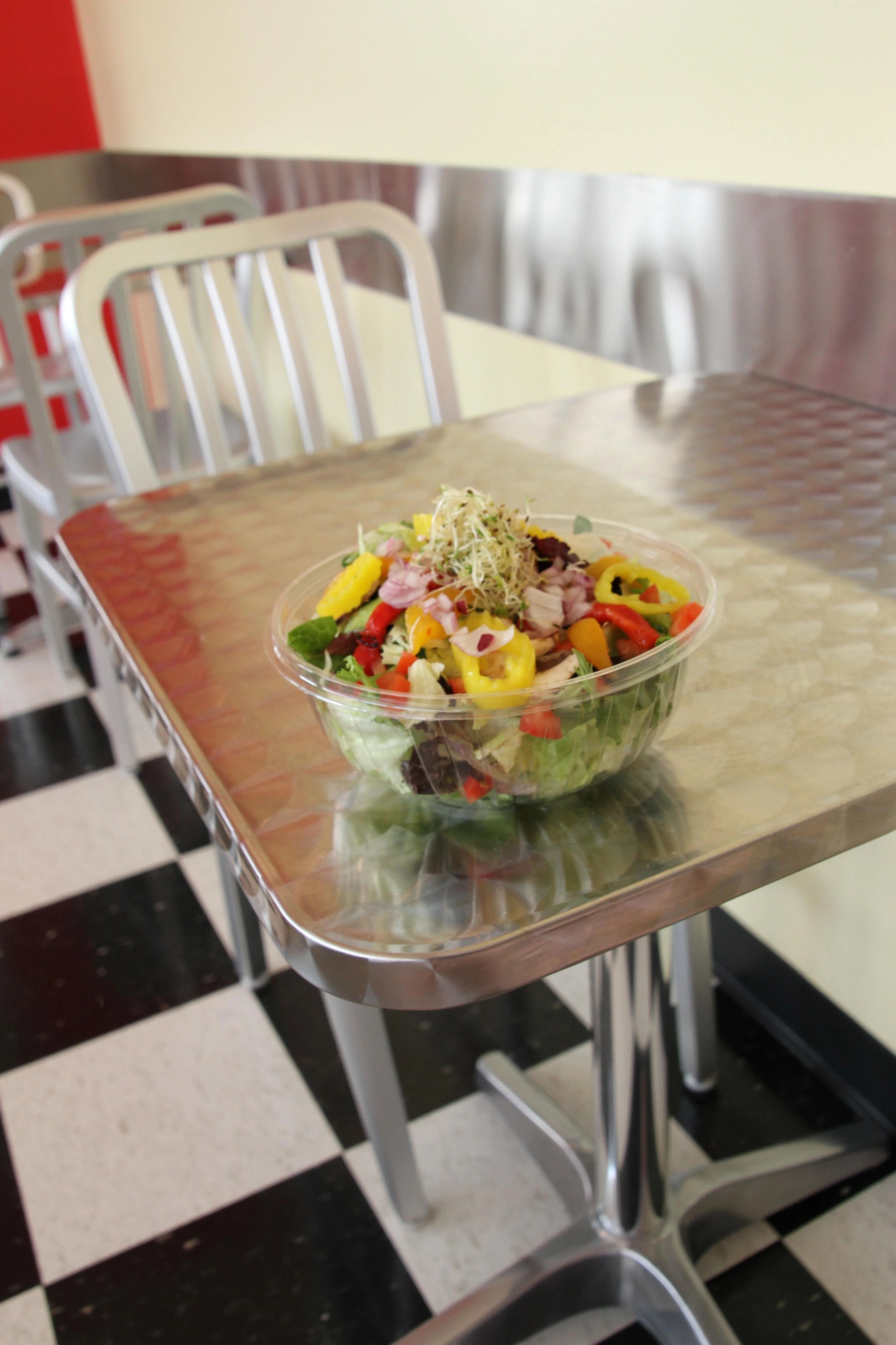 Fresh garden salad in a clear plastic bowl placed on a metal table inside a casual diner with checkered floors and aluminum chairs.