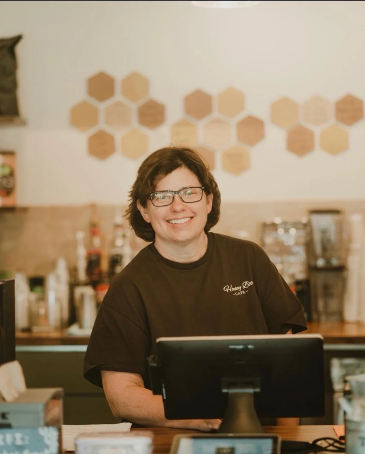 Smiling barista at the counter of a cozy café with hexagonal wall decor.