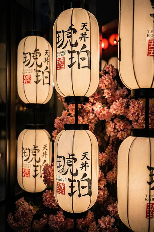 Glowing white Japanese paper lanterns featuring black brush calligraphy and red logo stamps. The lanterns are decoratively framed by a background of blooming pink cherry blossoms, capturing the restaurant's traditional aesthetic.
