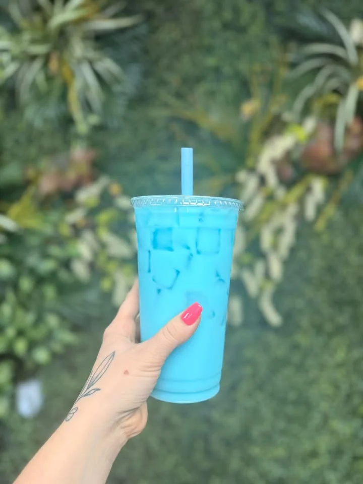 A vibrant blue beverage filled with ice, held against a backdrop of greenery.
