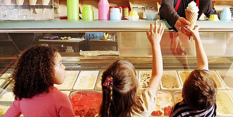 Children excitedly reaching for ice cream cones at a dessert shop