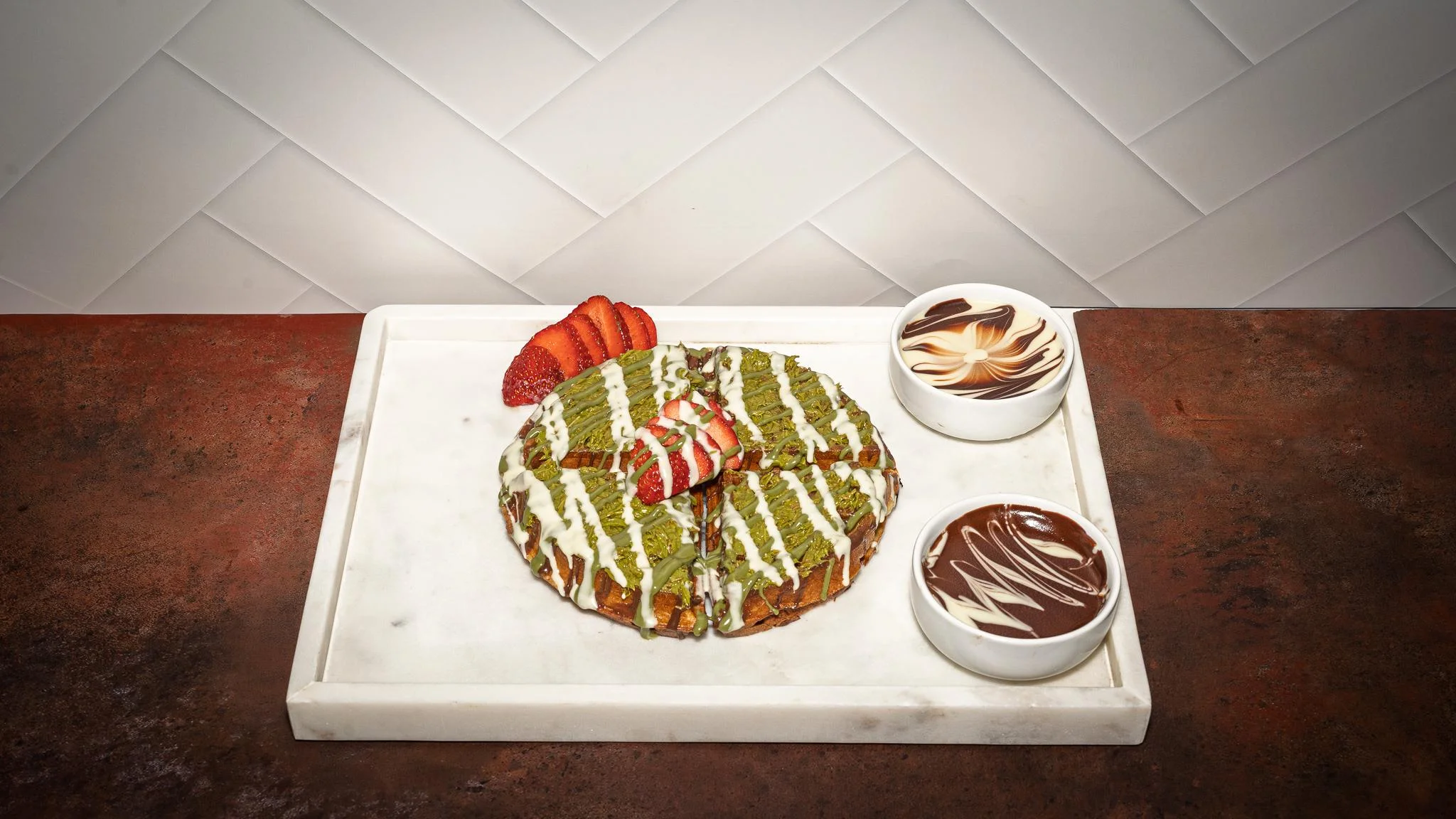 A round matcha-flavored cake topped with strawberries and drizzled with white icing, served with two small bowls of chocolate sauce.