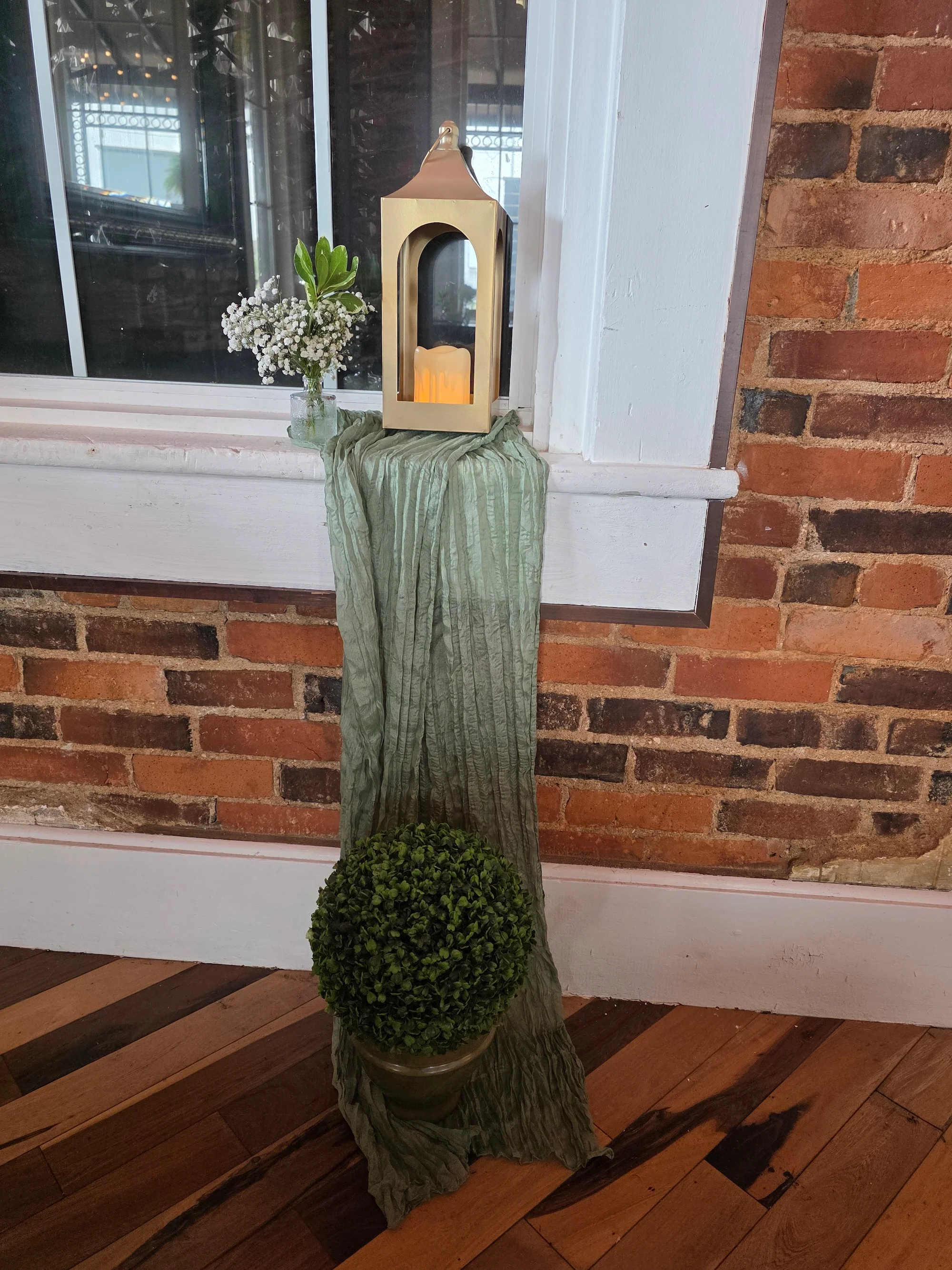 A decorative corner featuring a golden lantern, small vase with flowers, and a round potted plant, all set against a brick wall.