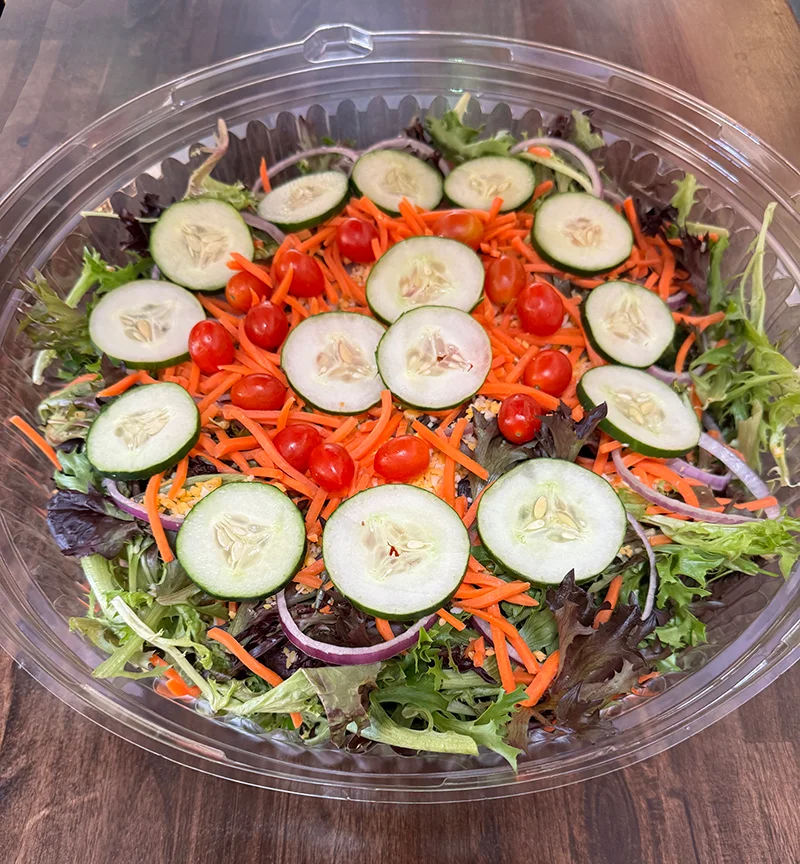 A fresh salad in a clear plastic container, featuring mixed greens, sliced cucumbers, cherry tomatoes, shredded carrots, and thinly sliced red onions, placed on a wooden table.