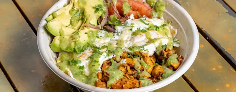 A bowl filled with layers of ingredients including avocado, meat, diced tomatoes, and topped with green sauce.